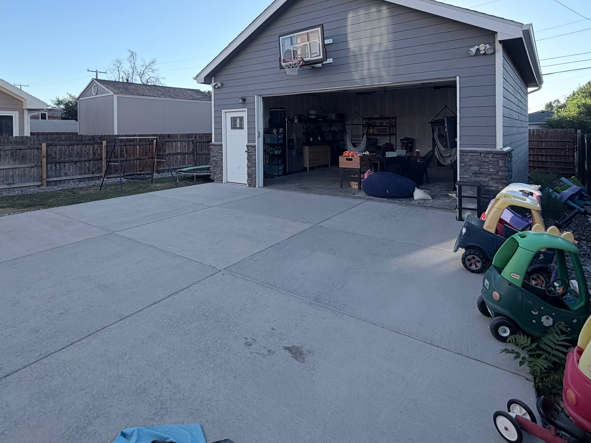 The detached garage from the backyard — wide-open doors reveal the indoor play space, with a basketball hoop above and ride-on toys nearby
