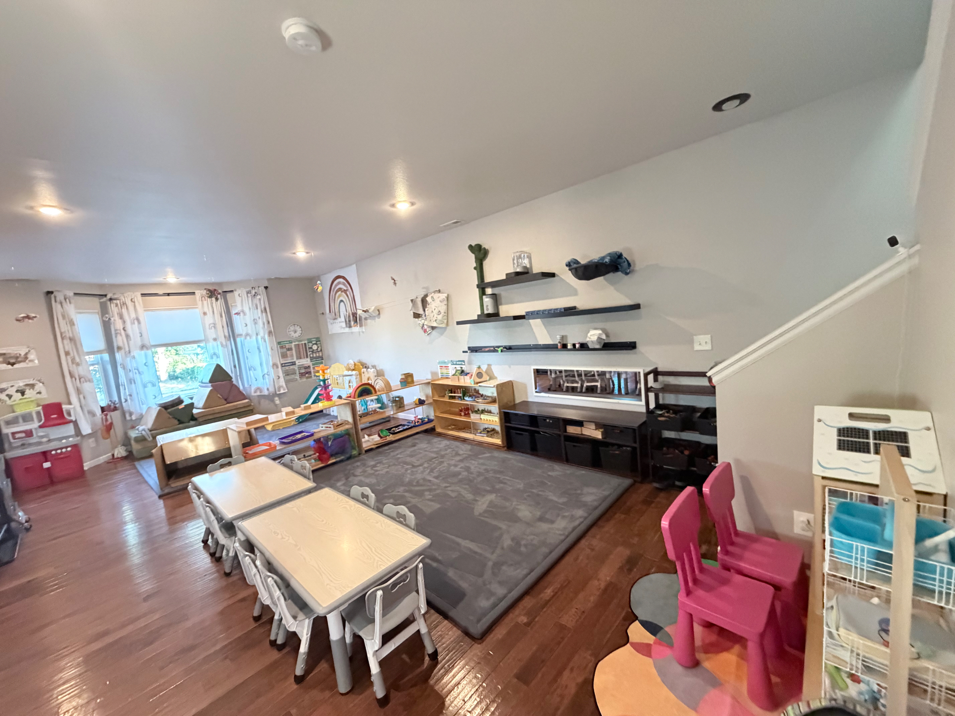 Wide view of the Montessori classroom with child-sized tables, low shelves, and a soft floor mat