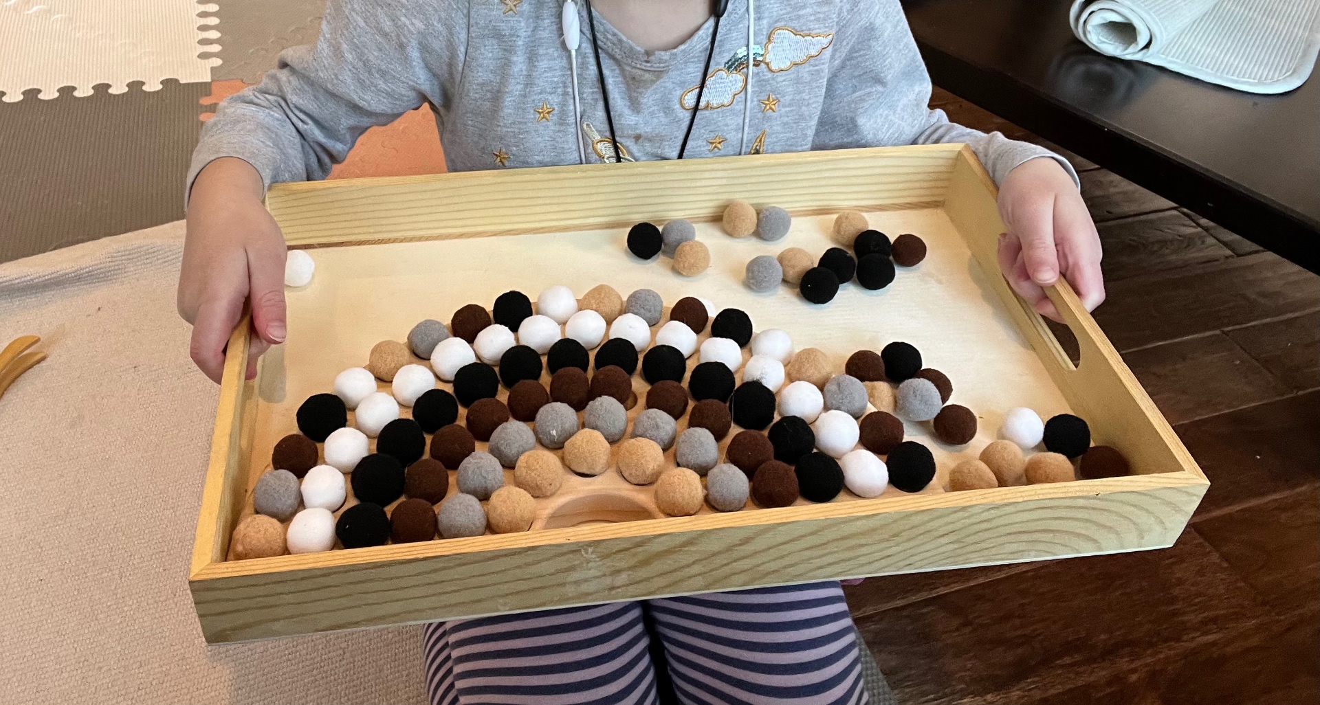 Child sorting neutral-toned felt balls on a wooden tray