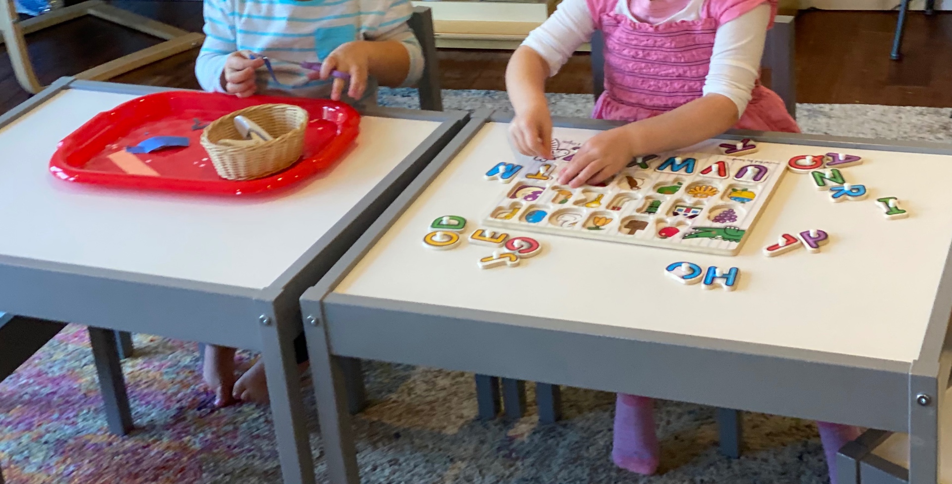 Two children working side-by-side at child-sized tables with a wooden alphabet puzzle